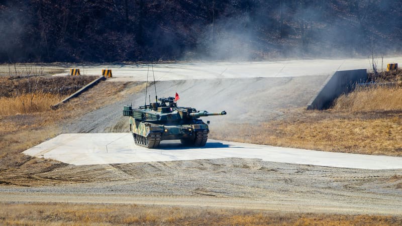 A South Korean K1-series tank with a red flag drives onto a concrete firing pad on a dusty training range