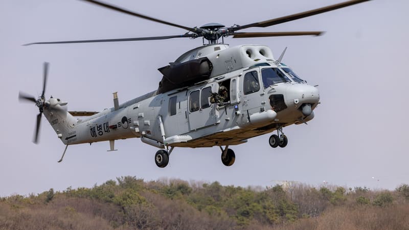 A grey South Korean Marine Corps MUH-1 Marineon helicopter with a door gunner descends to land over scrubland.