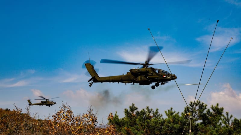 A U.S. Army AH-64E Apache flies low over autumn forest in South Korea, with a second Apache in the distance.