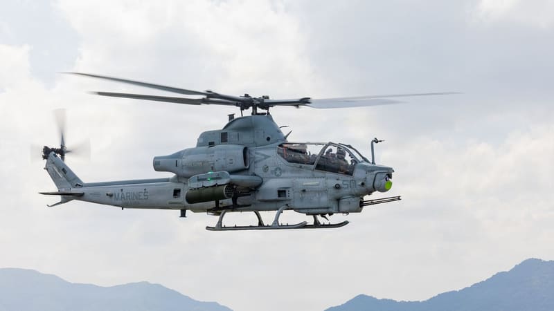 A U.S. Marine Corps AH-1Z Viper attack helicopter flies in profile over hazy mountains, nose turret gun visible.