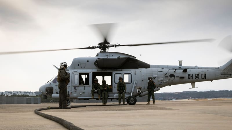 South Korean and U.S. Marines stand beside a grey Marineon helicopter on an airfield as a fuel hose runs across the ground.
