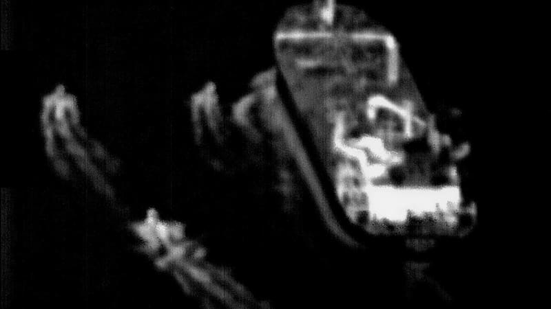 Grainy black-and-white aerial frame of three small fast boats approaching a large merchant ship