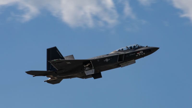 A dark grey KF-21 Boramae prototype fighter climbing with its landing gear retracting against a blue sky