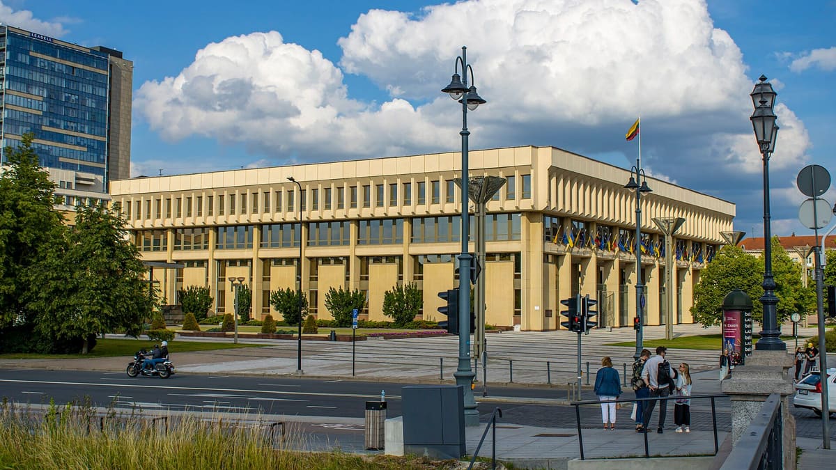 Seimas parliament building in Vilnius under a blue sky with large clouds, a Lithuanian flag on a pole above the roofline