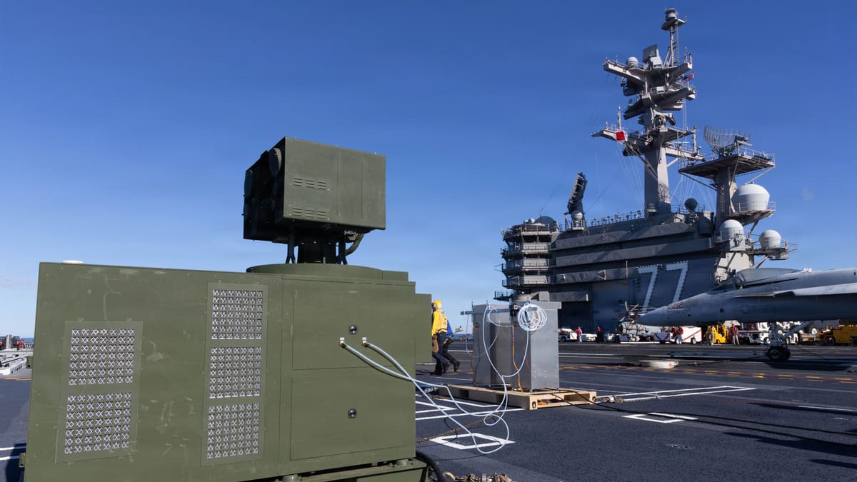 A green containerised LOCUST laser weapon on an aircraft carrier flight deck, with the carrier's island behind it