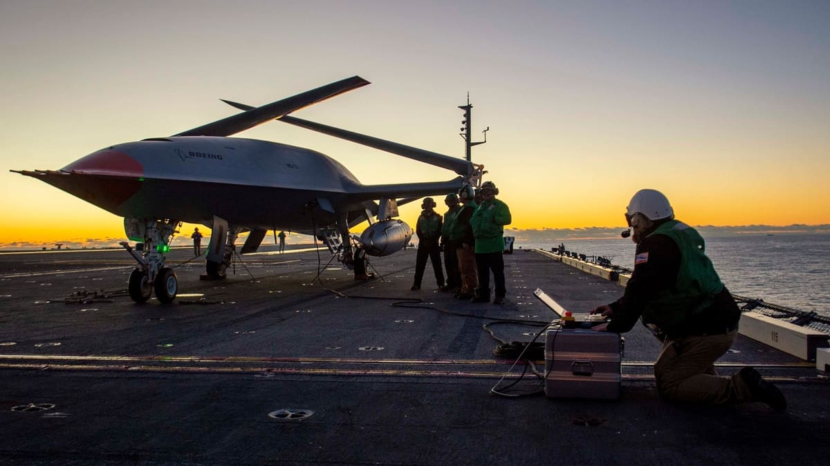 Sailor kneeling at a portable control unit beside an MQ-25 drone on a carrier deck against an orange horizon