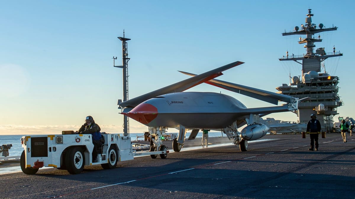 Tow tractor pulling an MQ-25 drone with folded wings across a carrier flight deck