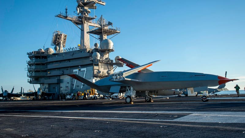 Grey MQ-25 tanker drone with red-tipped wings parked on a carrier deck beneath the island of USS George H.W. Bush