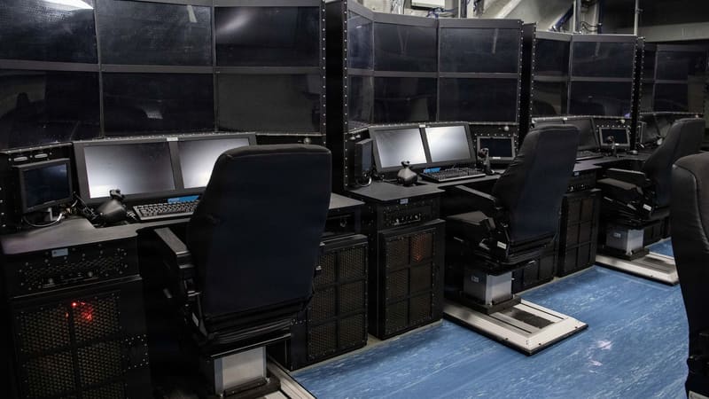 Rows of control consoles with monitors, keyboards and black seats in a compartment aboard an aircraft carrier