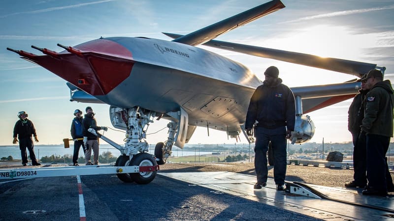 Sailors and Boeing staff stand beside the nose gear of a grey and red MQ-25 drone on a carrier deck in port