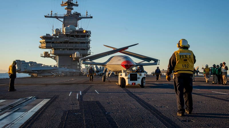 MQ-25 drone with wings folded towed by a tractor across a carrier deck in low sunlight as deck crew watch