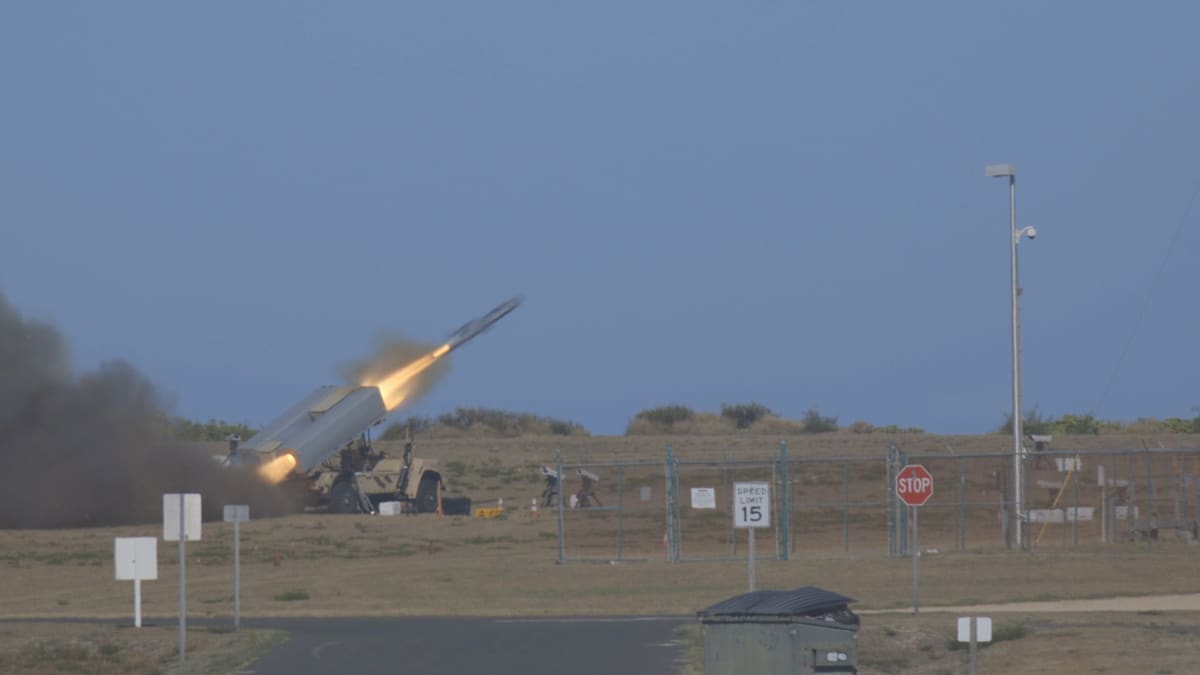 A missile leaves a box launcher on a military truck in a plume of smoke at a fenced range.