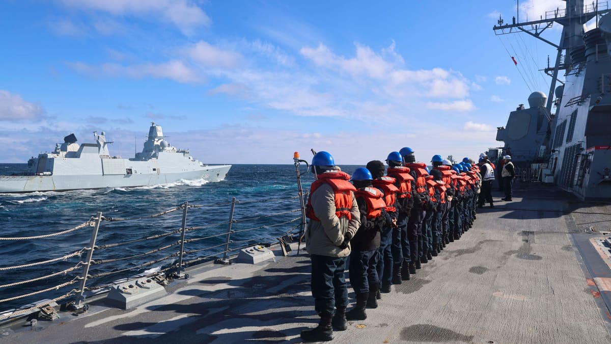 A grey Danish frigate sails past a line of U.S. sailors in life vests on a destroyer's deck.