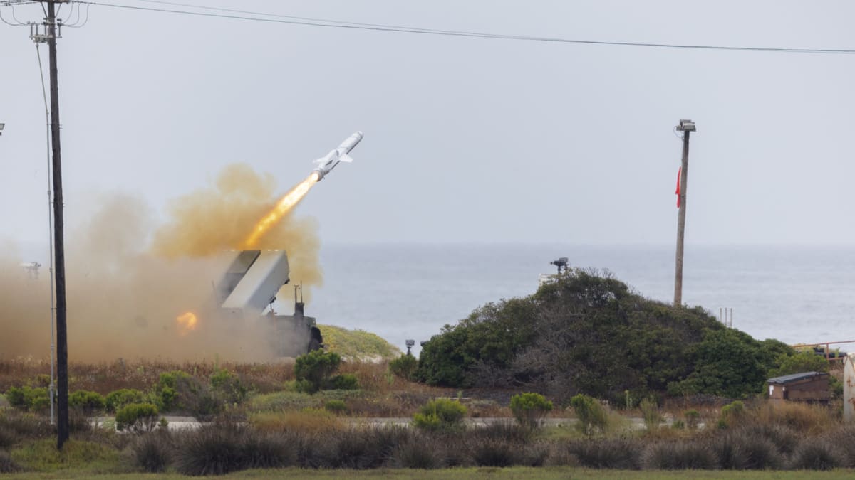 A Naval Strike Missile rises from a box launcher on a military truck in a cloud of dust beside the Pacific coast.