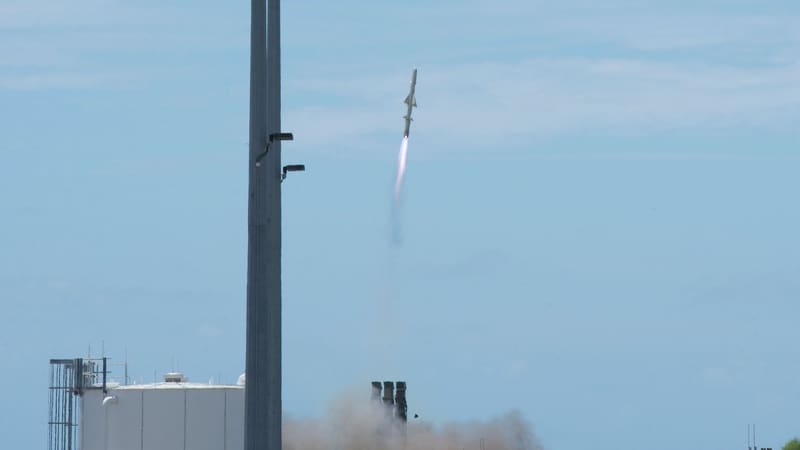 A missile climbs into a blue sky trailing smoke from a coastal launch site with a white tank and tall masts