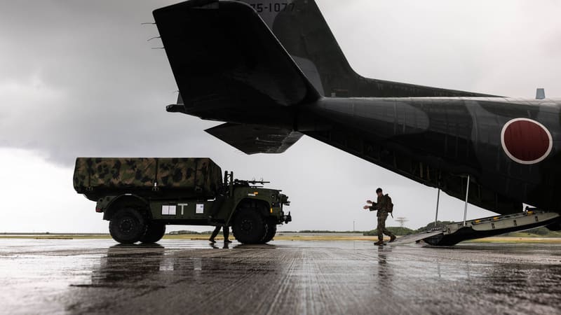 A U.S. Marine guides a tarpaulin-covered military truck towards the open ramp of a Japanese C-130 on a wet runway