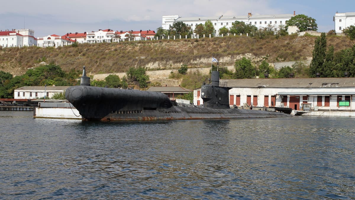 A black Soviet-built Romeo class submarine with a bulbous bow, moored in Sevastopol harbour below white hillside buildings.