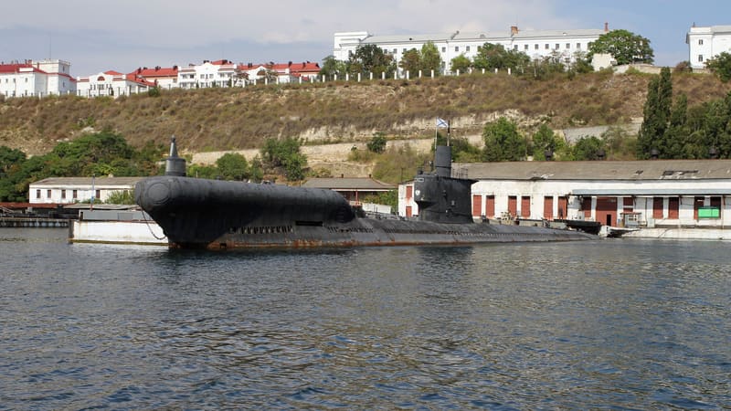 A black Soviet-built Romeo class submarine with a bulbous bow, moored in Sevastopol harbour below white hillside buildings.
