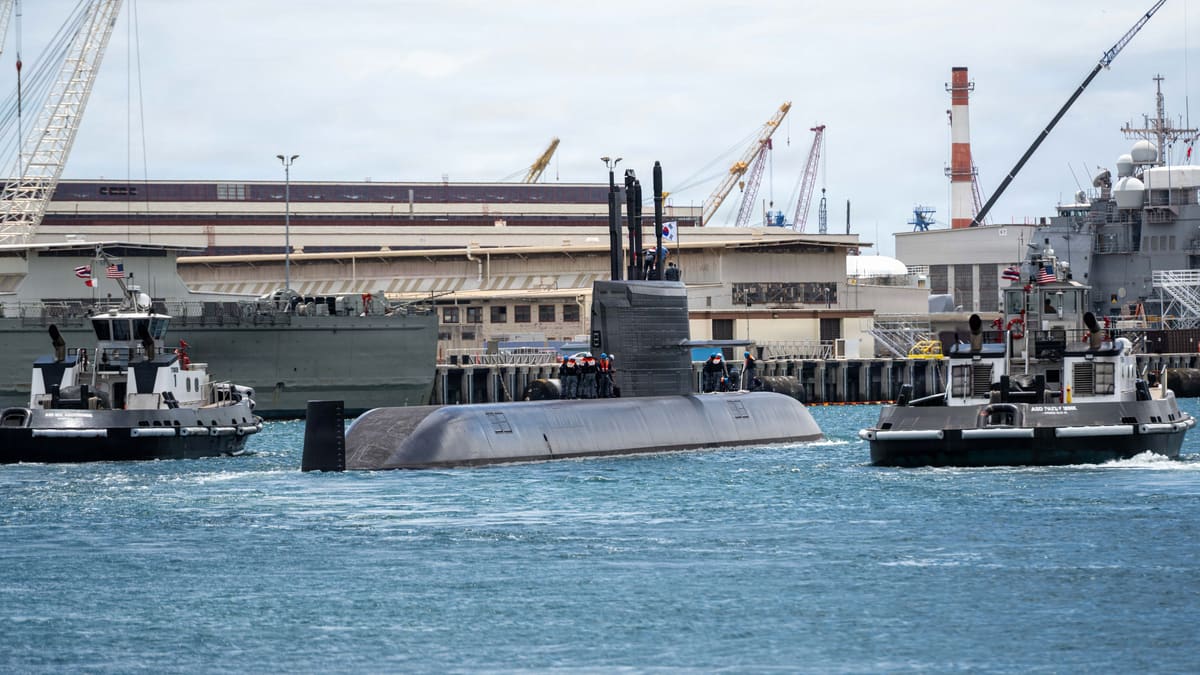 South Korean submarine ROKS Dosan Ahn Changho on the surface, escorted by two tugs in a harbour with cranes behind.