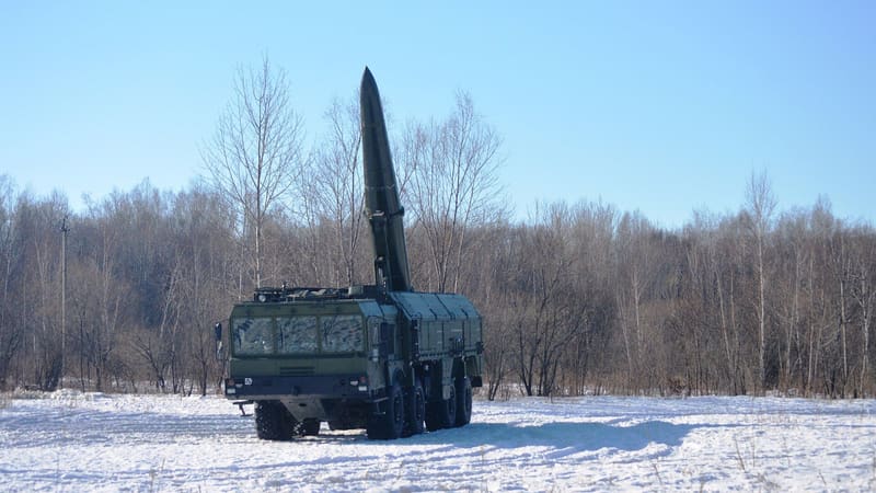 A Russian Iskander-M launcher with its ballistic missile raised, parked on snow in front of bare birch trees