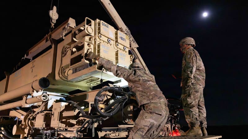 One U.S. soldier guides a training missile canister onto a Patriot launcher at night as another watches under a full moon
