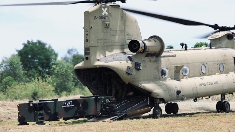 A U.S. Army CH-47 Chinook with its ramp down beside an inert Patriot missile training canister on a grass field
