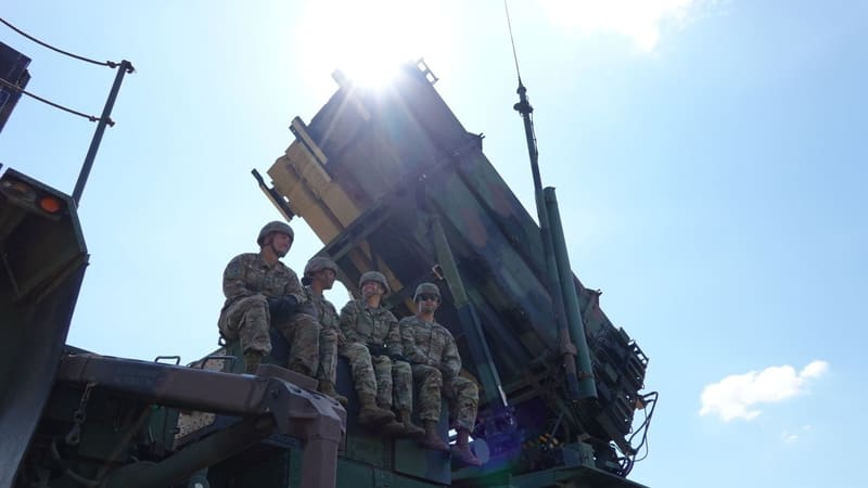 Four U.S. Army soldiers sit on a raised Patriot missile launcher against a bright blue sky