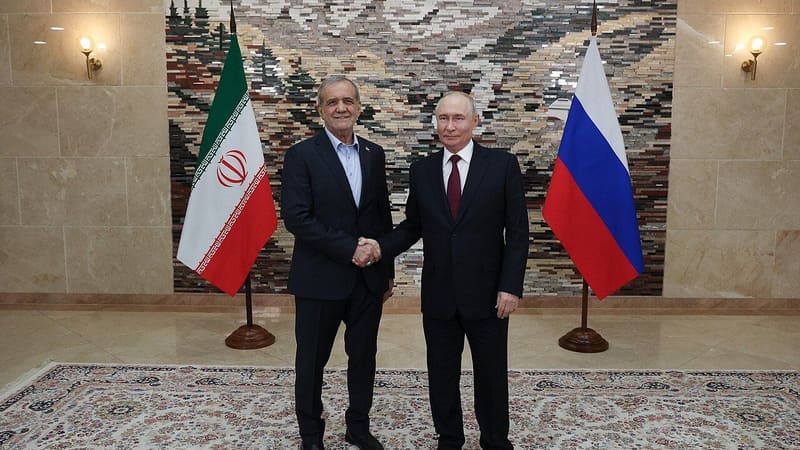 Two men in dark suits shake hands in front of an Iranian flag and a Russian flag in a marble hall