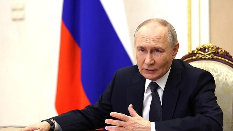 Vladimir Putin in a dark suit speaks at a table in front of the Russian flag