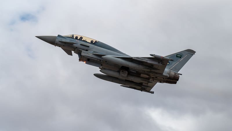 A two-seat Royal Saudi Air Force Typhoon with external fuel tanks climbs against a grey cloudy sky