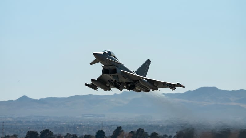 An RAF Typhoon FGR4 fighter climbs away from a desert runway with its landing gear retracting and mountains behind
