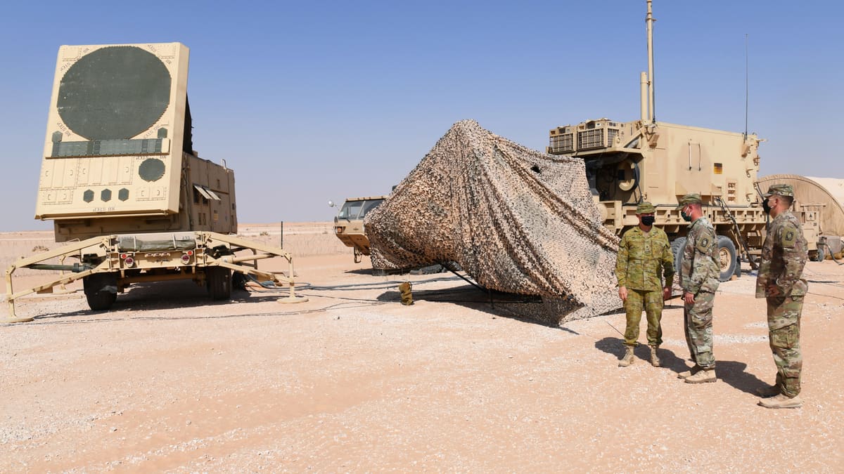Tan Patriot radar on a trailer in the desert beside a camouflage-netted vehicle, with soldiers standing nearby