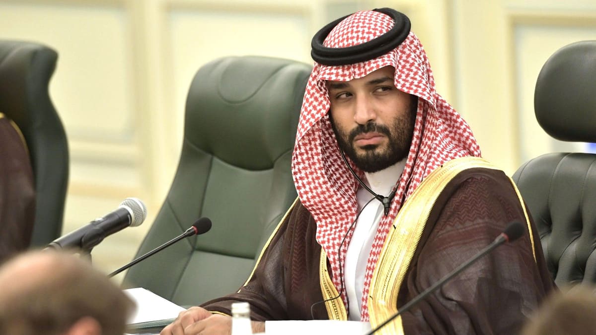 Saudi Crown Prince Mohammed bin Salman in a red-and-white checked headdress, seated at a conference table with microphones