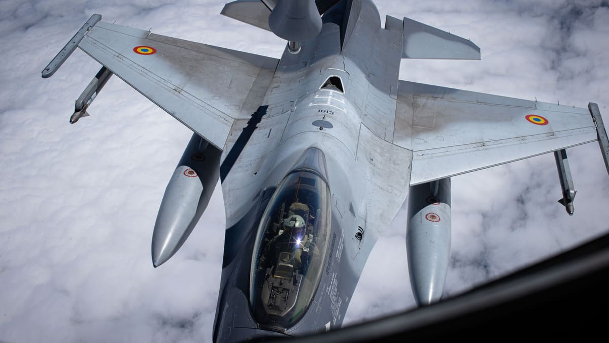 Romanian Air Force F-16 with red, yellow and blue roundels seen from above beneath a tanker's refuelling boom, clouds below