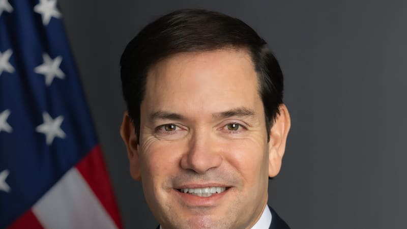 Official portrait of Marco Rubio in a dark suit in front of a US flag