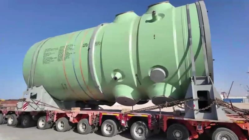 A large green cylindrical vessel with nozzles, chained to a red multi-axle transporter on a quay under a blue sky