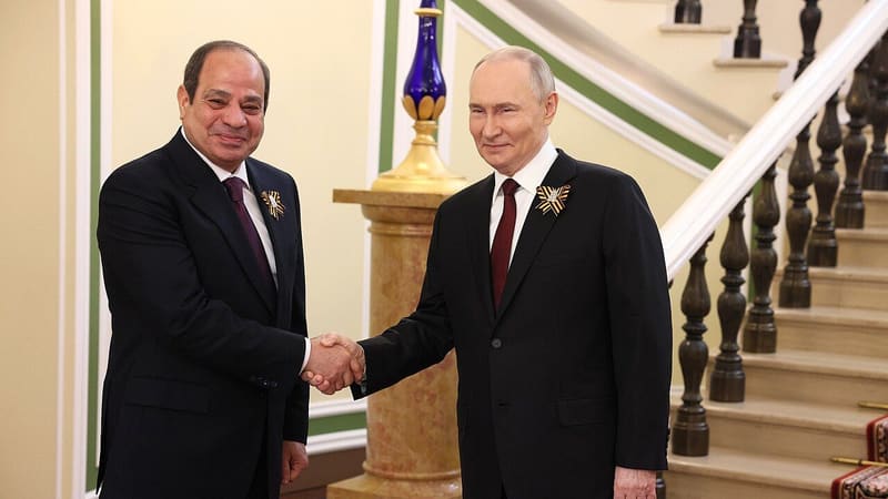 Two men in dark suits shake hands in front of a staircase, each wearing a striped ribbon pin on his lapel