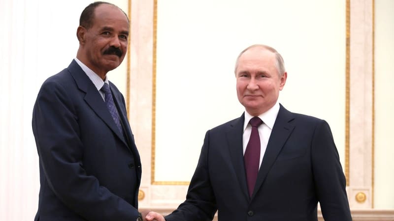Two men in dark suits shake hands in front of a cream wall with gold trim