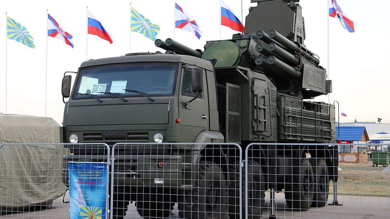 Pantsir-S1 vehicle with radar and missile tubes behind a fence, Russian and Air Force flags flying behind it