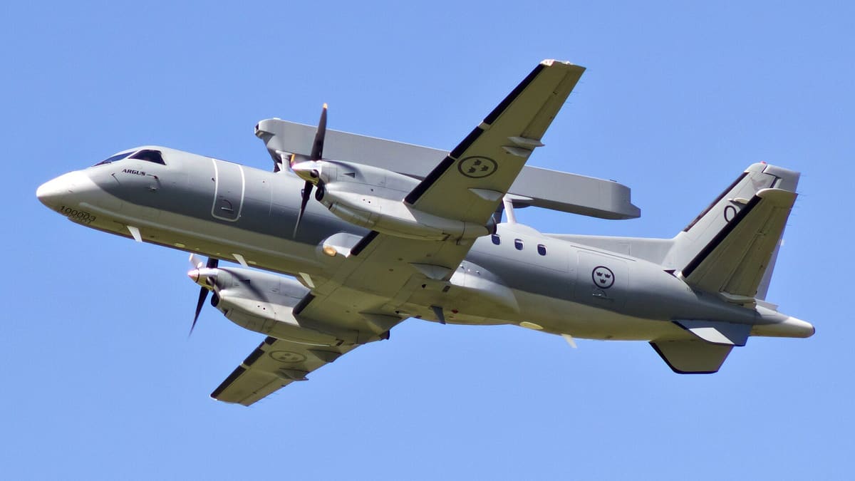 Swedish Air Force Saab 340 twin-propeller plane with a long radar on its back, seen from below climbing in blue sky