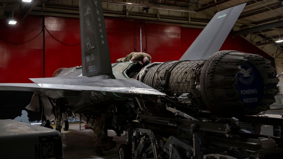 An F135 jet engine on a trailer is guided into the rear fuselage of an F-35A inside a hangar