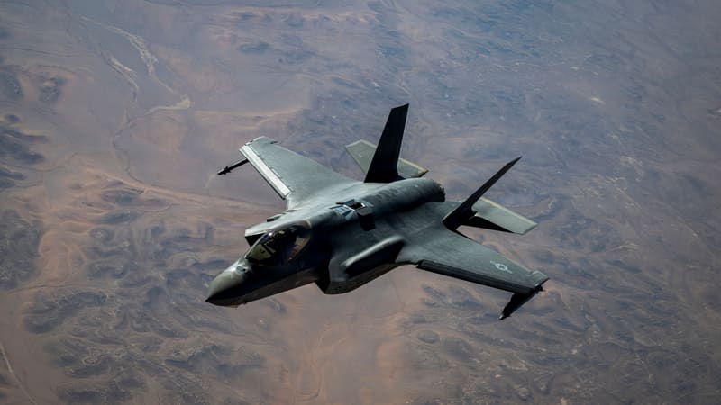 A grey U.S. Air Force F-35A stealth fighter banks over brown desert terrain seen from above