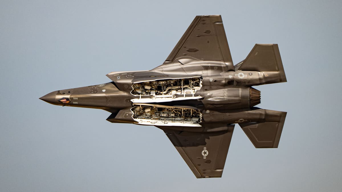 An F-35A seen from below in flight with both internal weapons bays open against a grey sky