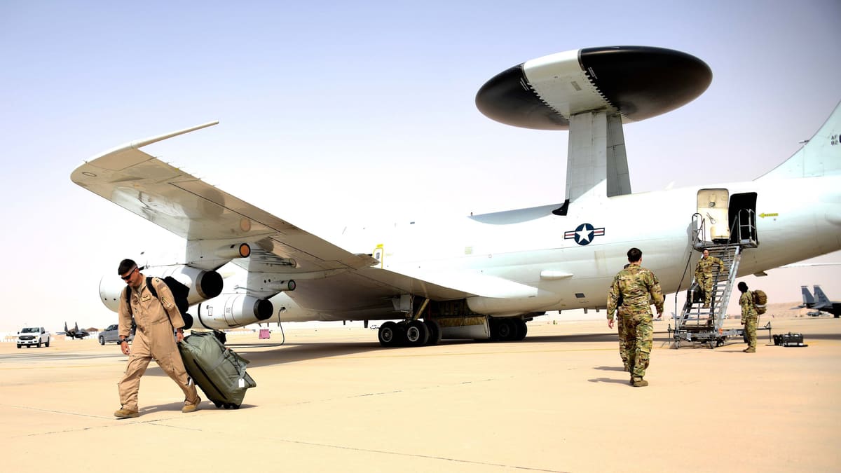 U.S. Air Force E-3G Sentry with its large radar dome parked on a desert apron as aircrew carry bags to and from it