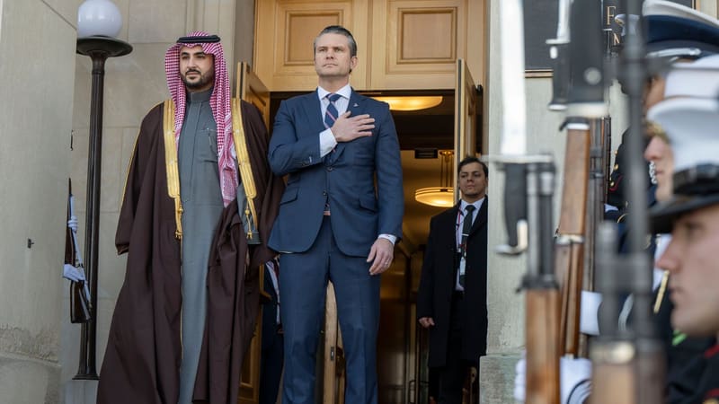 Prince Khalid bin Salman in keffiyeh and gold-trimmed robe beside Pete Hegseth, hand on heart, behind blurred guard rifles