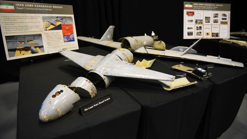 Damaged grey Qasef-1 drone wreckage on black display tables beside placards and an On Loan from Saudi Arabia sign