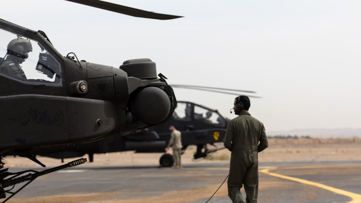 Apache nose and sensor turret with a helmeted crewman in the cockpit; a crewman in a flight suit walks toward a second Apache