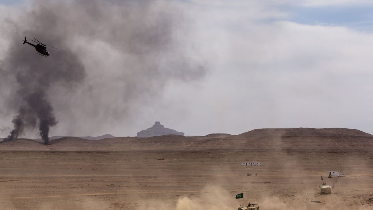 A light scout helicopter flies past columns of black smoke above a desert range as Humvees with Saudi flags drive below