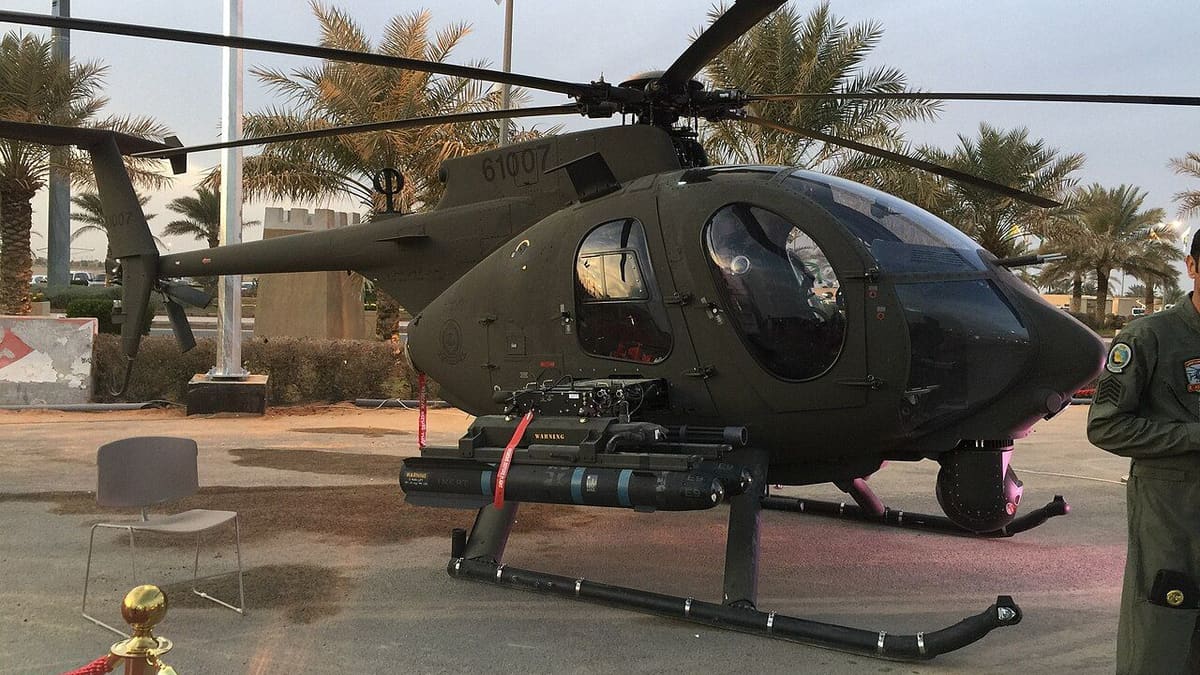 Dark green AH-6i light helicopter with a Hellfire missile, minigun pod and chin sensor turret, parked under palm trees