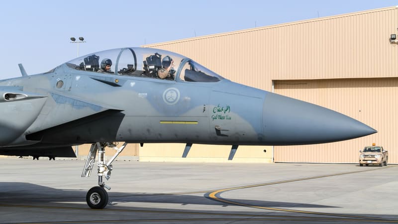 Two-seat Royal Saudi Air Force F-15 with RSAF roundel taxiing past a tan hangar, front-seat crew member waving